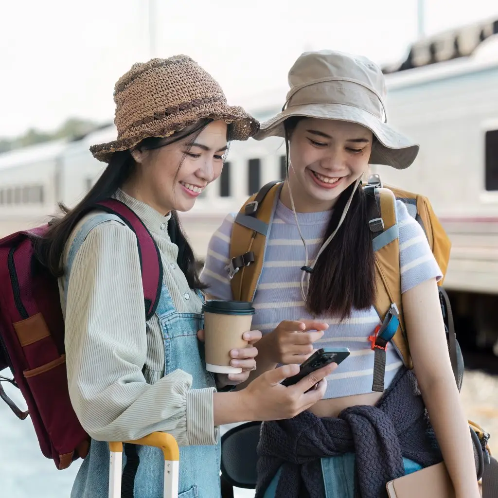 Two young women with backpacks and hats at a train station, smiling and looking at a phone, ready for their travel adventure.