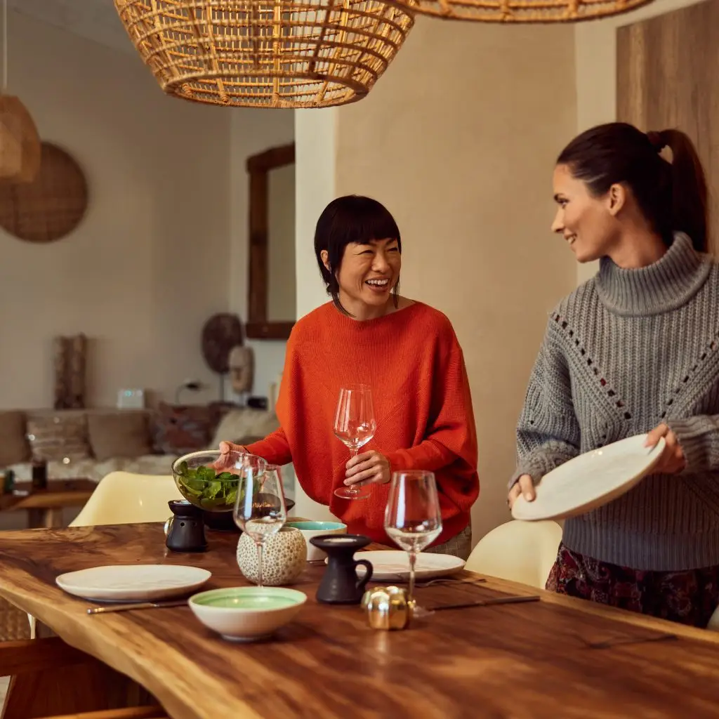 Smiling female friends looking at each other, serving a table for dinner, at home.